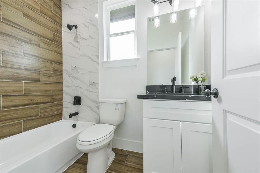 Modern bathroom featuring a combination of wood-like tile and marble-style walls, a white bathtub, toilet, and vanity with a black countertop and integrated sink. It has a frosted glass window and stylish overhead lighting above the mirror.