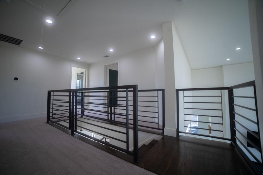 The upper-level landing serves as a stunning gallery space, featuring sleek horizontal iron railings that mirror the home’s modern entrance. This open design ensures uninterrupted sightlines to the living areas below, maintaining a sense of light and airiness throughout the second floor