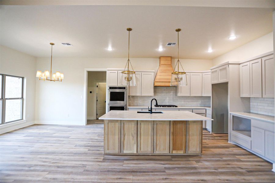 Kitchen with a center island with sink, two tone cabinetry, stainless steel double oven, hanging lights, and light wood-style flooring