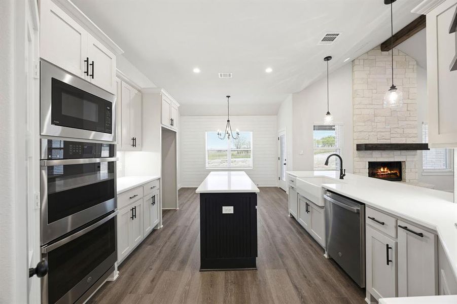 Modern kitchen featuring white cabinetry with black hardware, stainless steel appliances, a center island with a dark base, wood-finish flooring, and a stone fireplace with a substantial wood mantel