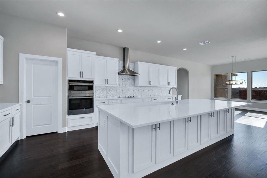 Kitchen featuring decorative backsplash, wall chimney exhaust hood, dark hardwood / wood-style floors, double oven, and a kitchen island with sink