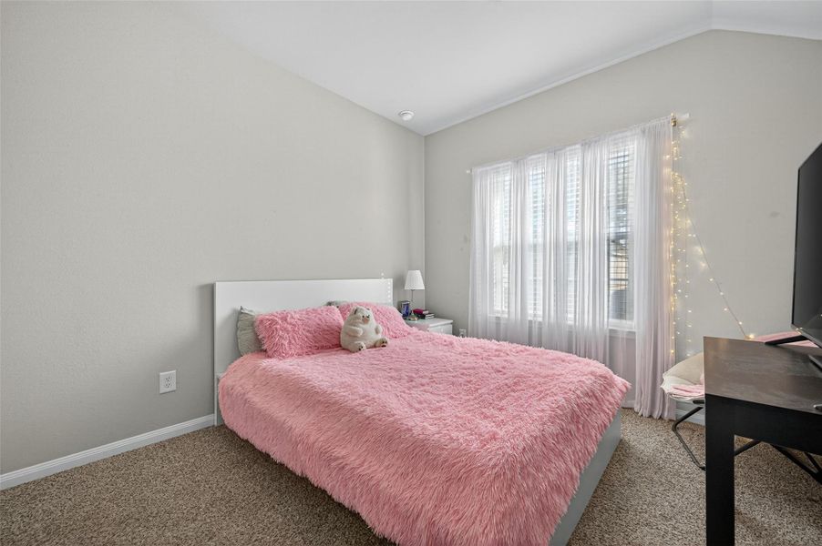 Cozy bedroom with neutral walls, featuring a bed adorned with a pink fluffy comforter. The room is brightened by natural light from a large window with sheer curtains and string lights. Carpeted flooring and a small desk with a TV complete the space, offering a comfortable and inviting atmosphere.