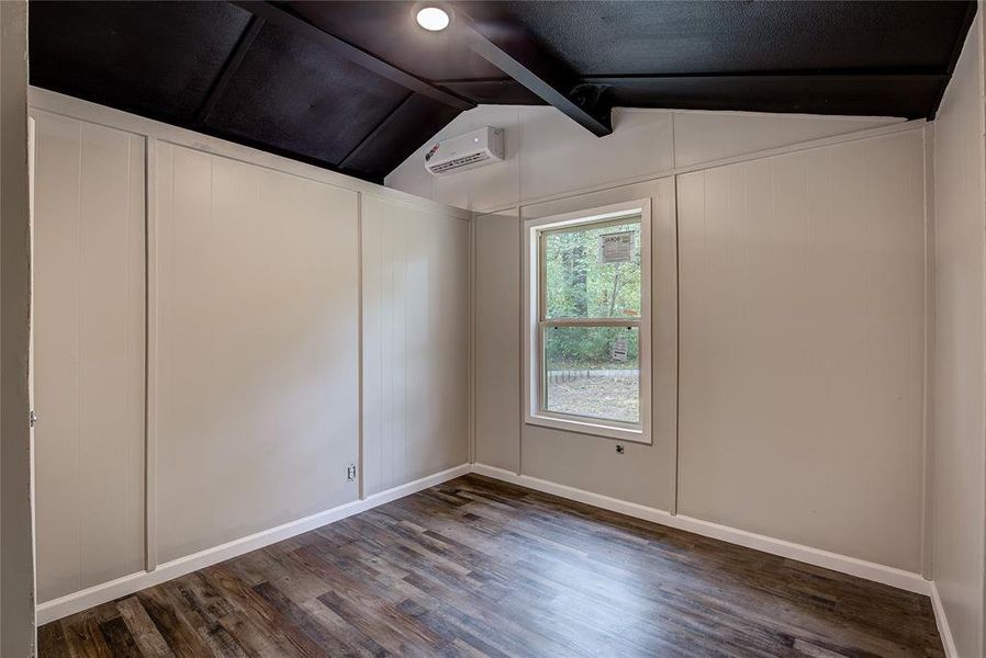 Empty room featuring dark wood-type flooring, a wall mounted air conditioner, and lofted ceiling with beams