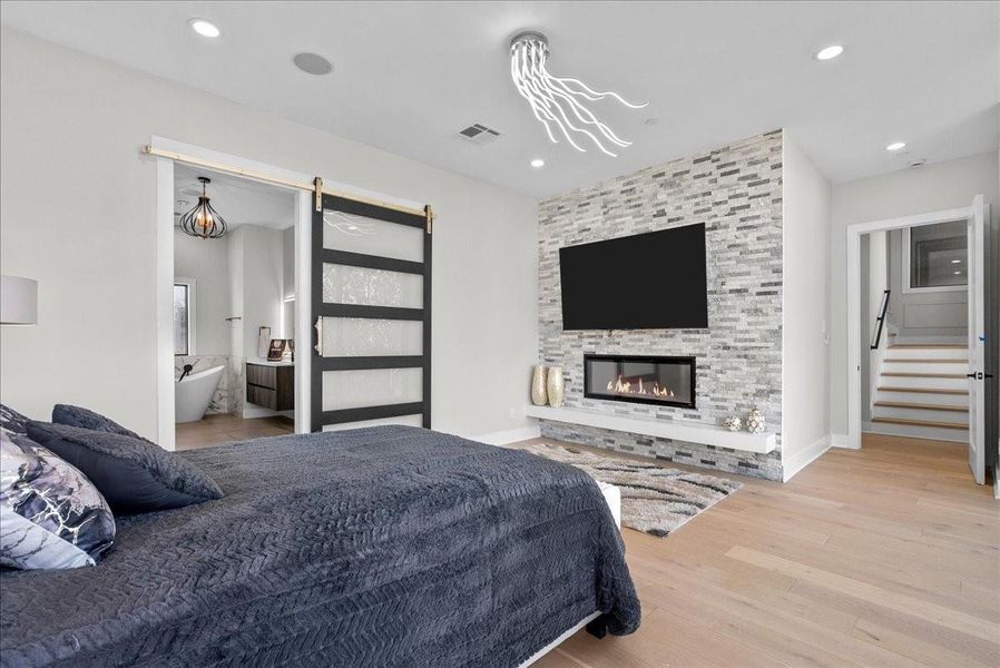 This modern bedroom features a cozy fireplace with a stone accent wall, a mounted TV, and sleek wood flooring. A sliding barn door leads to a luxurious bathroom with a soaking tub. Recessed lighting and a contemporary ceiling fixture add elegance. This modern bedroom features a cozy fireplace with a stone accent wall, a mounted TV, and sleek wood flooring. A sliding barn door leads to a luxurious bathroom with a soaking tub. Recessed lighting and a contemporary ceiling fixture add elegance.