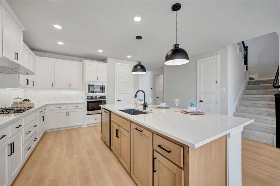 Kitchen with two tone cabinets, light wood-style flooring, decorative light fixtures, an island with sink, and backsplash