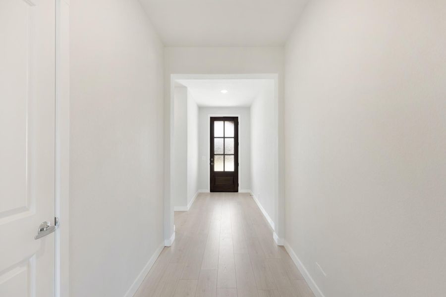 Narrow hallway with white walls, light wood floors, and dark wood front door with glass panels