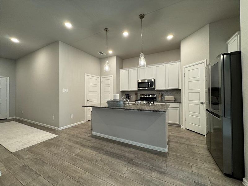 Modern kitchen featuring a central island with a dark countertop, white shaker-style cabinetry, stainless steel appliances, recessed lighting, and wood-finish flooring Modern kitchen featuring a central island with a dark countertop, white shaker-style cabinetry, stainless steel appliances, recessed lighting, and wood-finish flooring