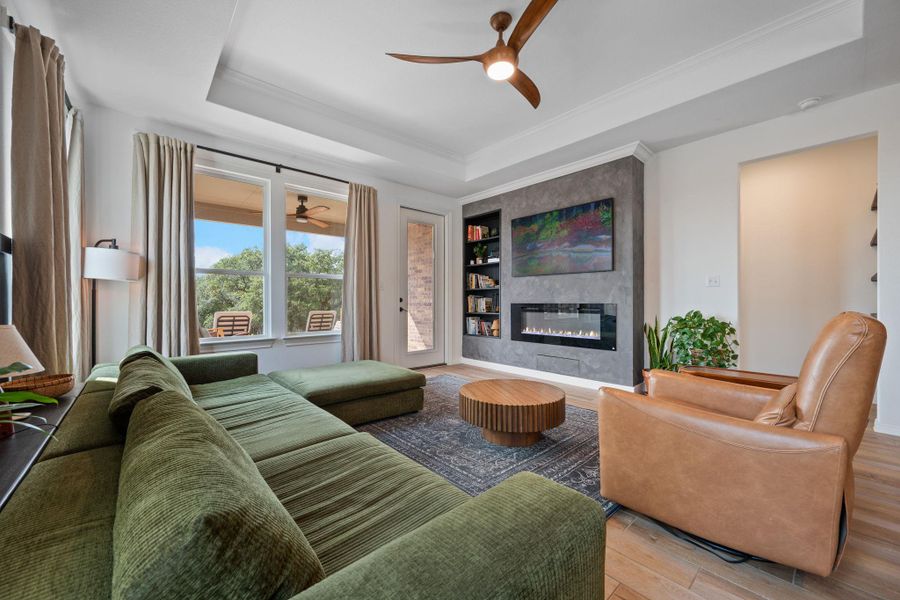 Living room featuring a raised ceiling, a ceiling fan, a large fireplace, wood finished floors, and crown molding Living room featuring a raised ceiling, a ceiling fan, a large fireplace, wood finished floors, and crown molding
