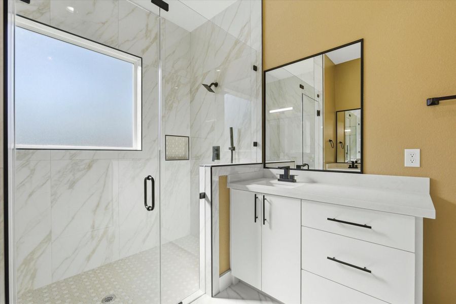 The primary shower features three shower heads, including a ceiling-mounted rain shower, a handheld, and a wall mounted fixture. Additional highlights include two built-in niches, a built-in bench, and beautiful hexagon tile flooring accented by large tile on the walls that matches the rest of the bathroom.