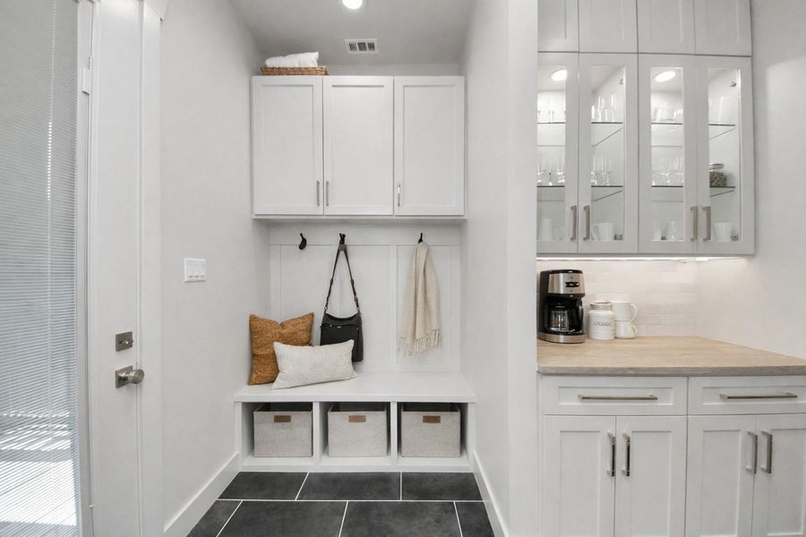 Thoughtfully designed for everyday convenience, this custom mud nook features built-in bench seating, upper cabinetry, storage cubbies, and decorative hooks, while the adjacent butler's pantry offers additional cabinetry, display shelving, and countertop workspace perfect for a coffee bar, beverage station, or extra serving area.