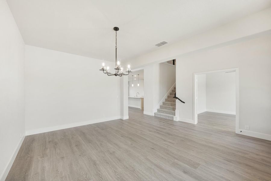 Open dining area with chandelier, light wood floors, white walls, and staircase visible in background.