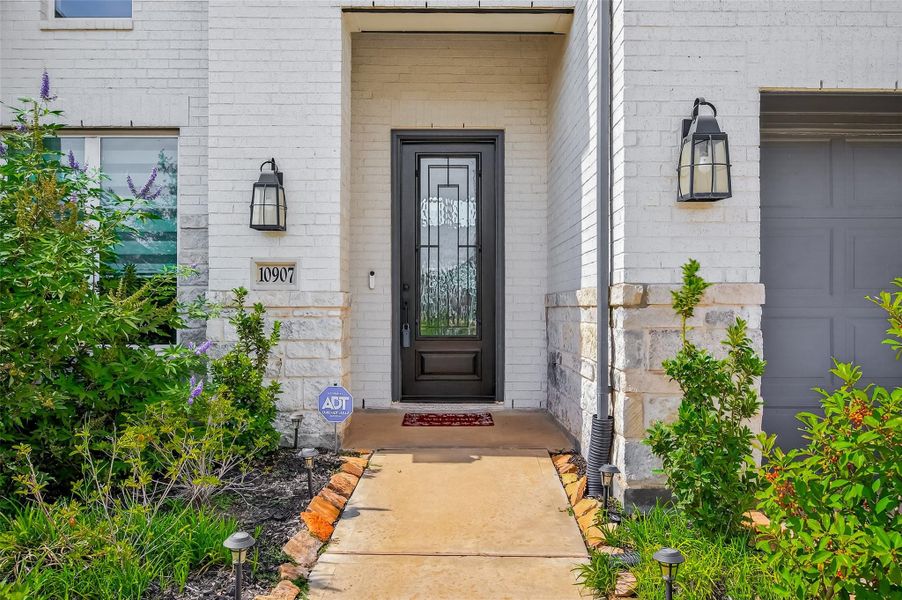 Charming front entry framed by lush native landscaping and warm stone accents. The glass-paneled door with wrought iron detail creates an elegant and secure first impression.