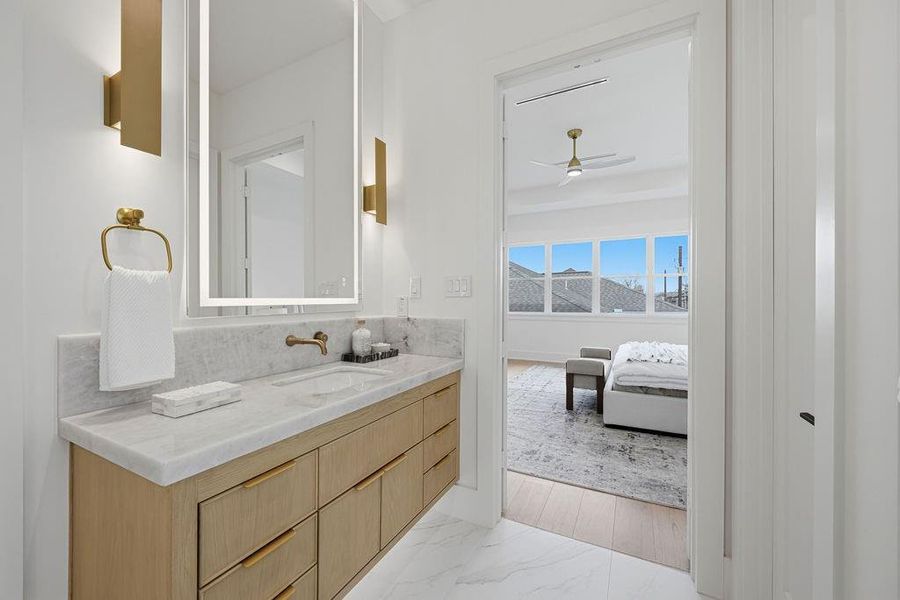 This sleek "his" station is anchored by a large, backlit designer mirror and contemporary gold sconces that provide a soft, ambient glow. Brushed gold hardware and a wall-mounted faucet maintain the home’s signature high-end aesthetic. The space offers a direct view into the light-filled primary bedroom, ensuring a seamless morning routine.