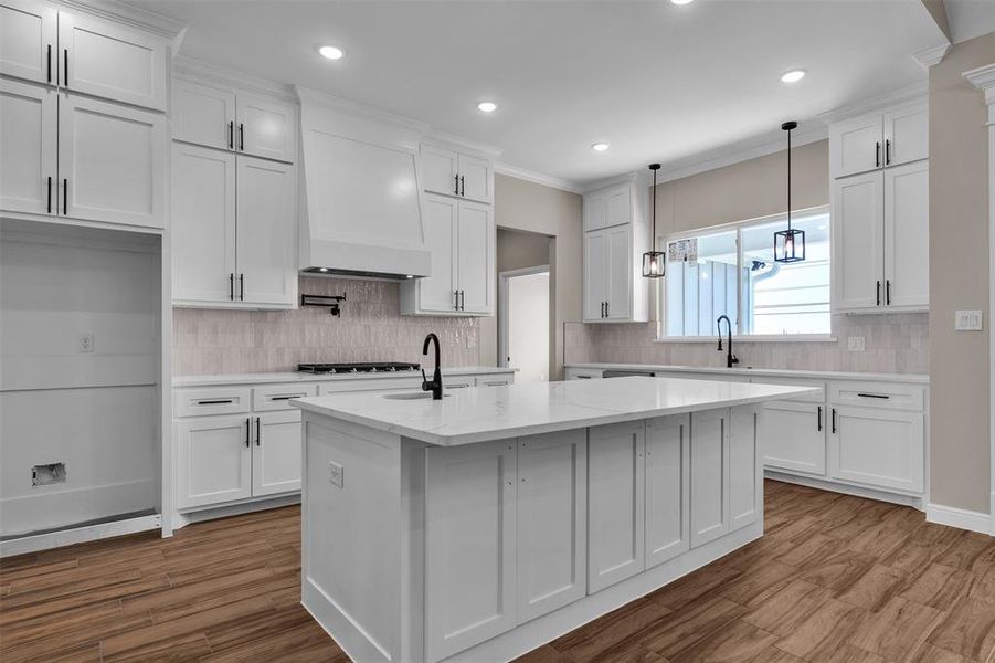 Kitchen with decorative backsplash, white cabinets, a center island with sink, ornamental molding, and pendant lighting Kitchen with decorative backsplash, white cabinets, a center island with sink, ornamental molding, and pendant lighting