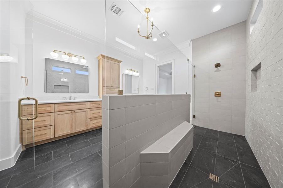 Spacious bathroom featuring a glass-enclosed shower with a built-in bench, dual vanity with natural wood-tone cabinetry, white countertop, and dark slate-style floor tiling