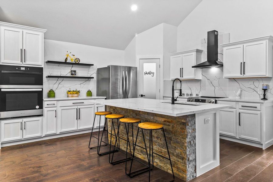 This modern kitchen features sleek white cabinetry, a spacious island with a stone accent, and stylish barstools. It includes stainless steel appliances, a double oven, and a chic range hood. The pantry door adds a functional touch, while open shelving provides decorative storage options.