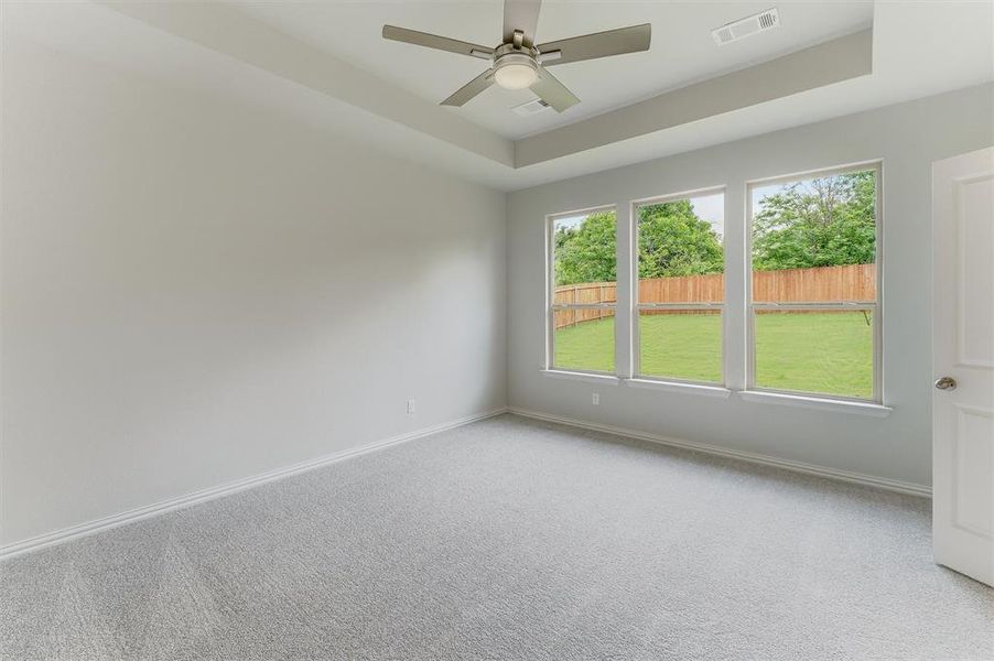 Spacious room featuring a tray ceiling with a contemporary ceiling fan, three large windows, light gray carpeting, and white baseboards