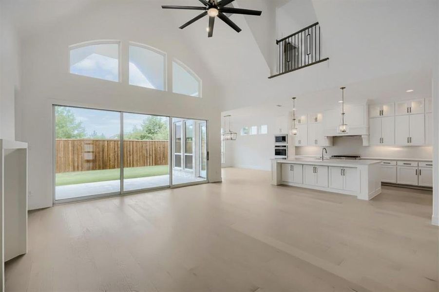 Vaulted ceiling great room featuring wood-finish flooring, a contemporary kitchen with white cabinetry and an island, a modern ceiling fan, and extensive sliding glass doors opening to a fenced backyard Vaulted ceiling great room featuring wood-finish flooring, a contemporary kitchen with white cabinetry and an island, a modern ceiling fan, and extensive sliding glass doors opening to a fenced backyard