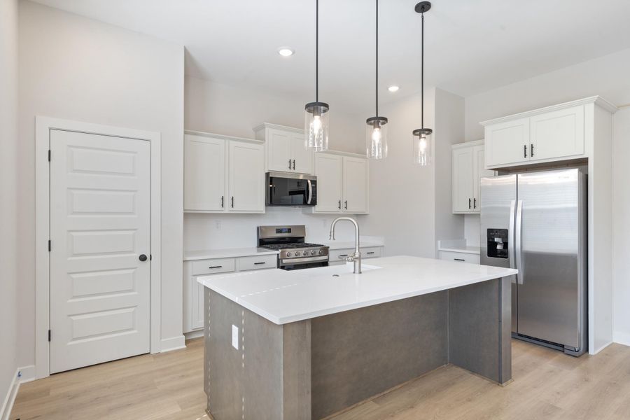 Kitchen featuring an island with a built-in sink, stainless steel appliances, white cabinetry, pendant lighting, and wood-finish flooring Kitchen featuring an island with a built-in sink, stainless steel appliances, white cabinetry, pendant lighting, and wood-finish flooring