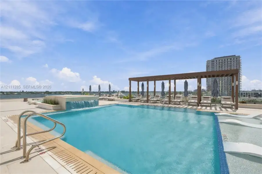 Rooftop swimming pool featuring a waterfall edge, integrated shallow sun shelf, metal handrails, and a covered pergola Rooftop swimming pool featuring a waterfall edge, integrated shallow sun shelf, metal handrails, and a covered pergola