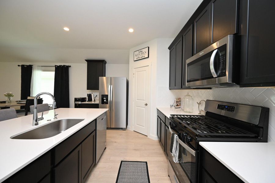 Modern kitchen with sleek dark cabinetry, stainless steel appliances, and white countertops. Features a large island with a sink and an open layout leading to a dining area. Modern kitchen with sleek dark cabinetry, stainless steel appliances, and white countertops. Features a large island with a sink and an open layout leading to a dining area.