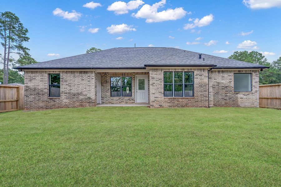 This photo shows the back of a single-story brick house with a spacious, well-maintained lawn. The home features a covered patio area, multiple windows, and a fenced backyard, ideal for privacy and outdoor activities.