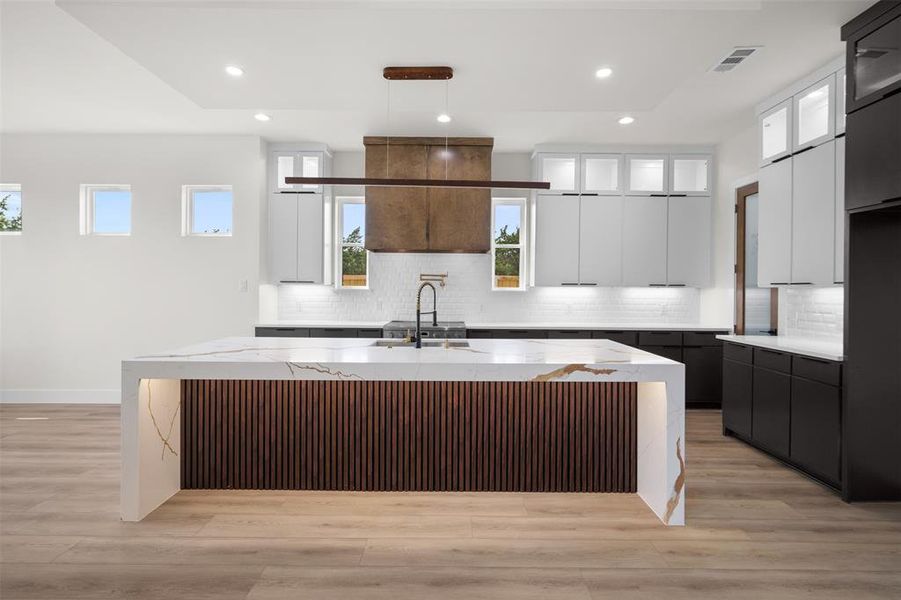 The kitchen features a large island with a waterfall countertop, a modern linear pendant light fixture, and two-tone cabinetry with contrasting finishes The kitchen features a large island with a waterfall countertop, a modern linear pendant light fixture, and two-tone cabinetry with contrasting finishes