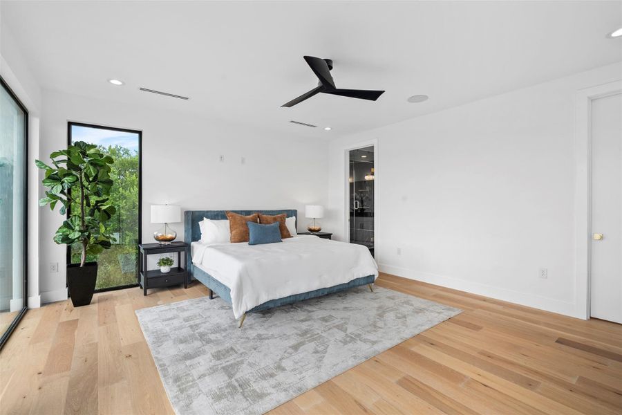 This bedroom features light hardwood floors, a large window providing views of greenery, and a modern ceiling fan