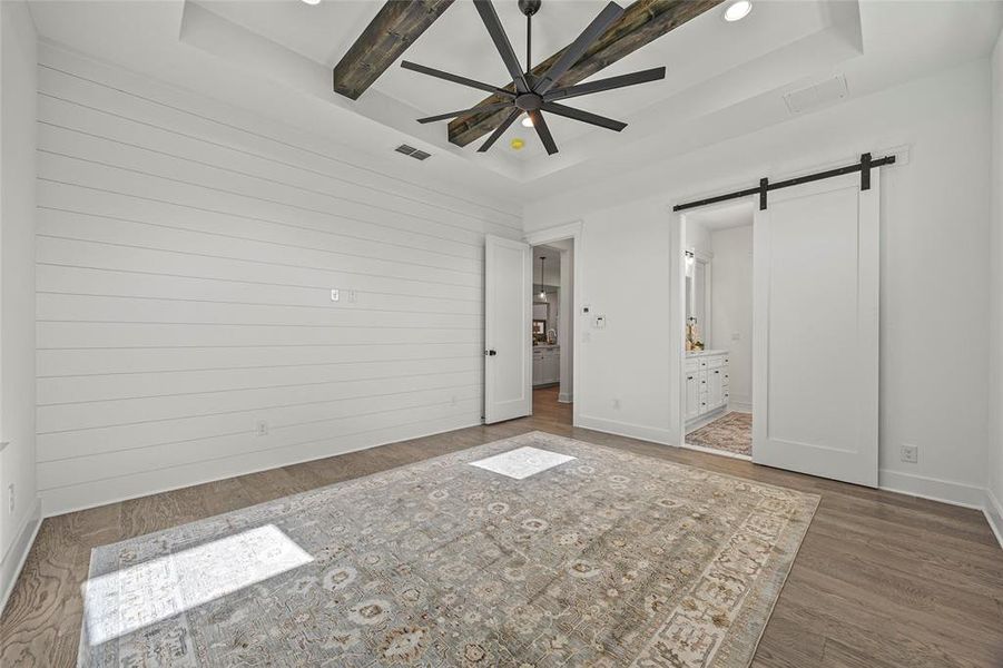 Unfurnished bedroom with ensuite bathroom, beamed ceiling, a barn door, hardwood / wood-style flooring, and a raised ceiling