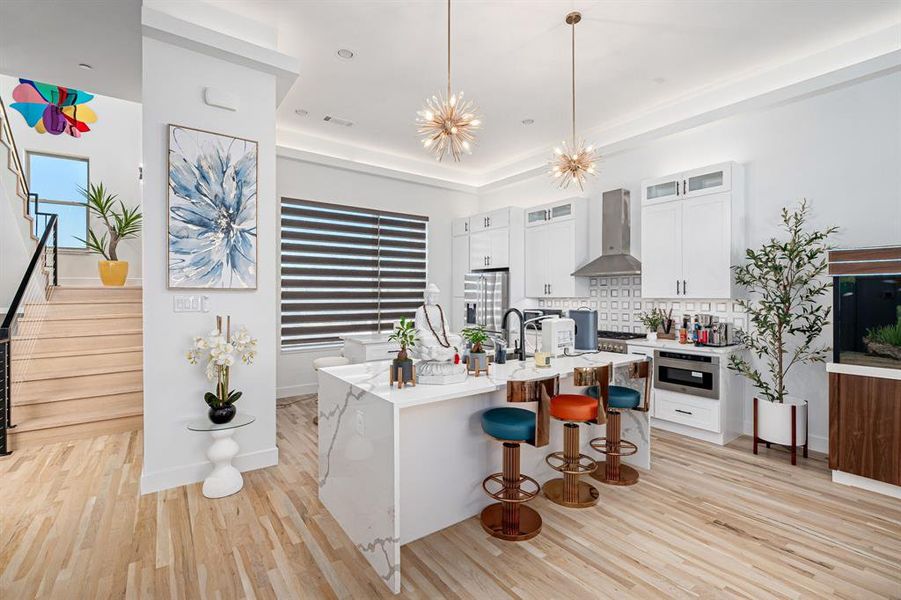 Kitchen with wall chimney range hood, tasteful backsplash, decorative light fixtures, white cabinetry, and a chandelier