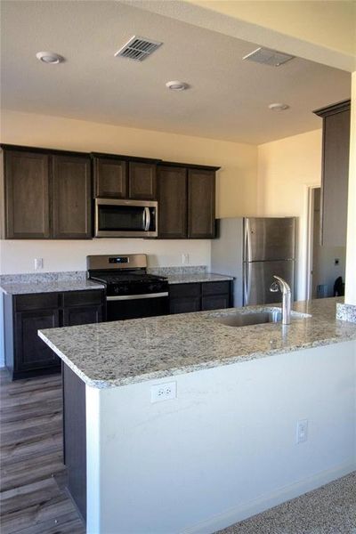 Kitchen with appliances with stainless steel finishes, dark brown cabinets, light stone countertops, and dark wood-type flooring