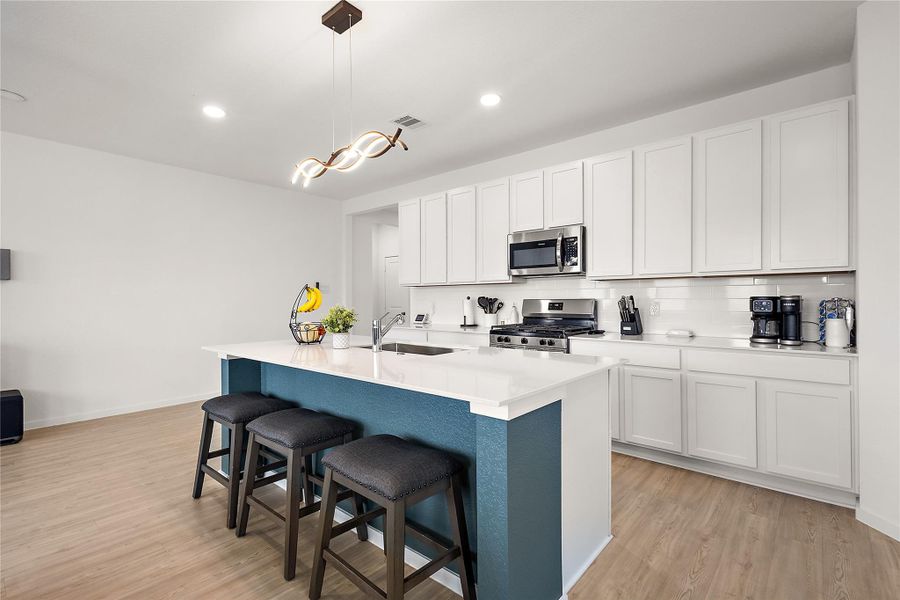 This modern kitchen features sleek white cabinetry, stainless steel appliances, and a spacious island with seating. The room is bright and open, complemented by stylish lighting and light wood flooring, creating an inviting space for cooking and gathering.