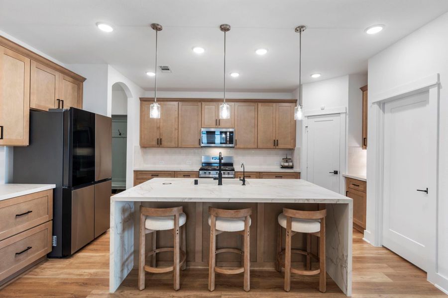 Modern kitchen features a spacious island with seating for three, sleek black appliances, and ample wooden cabinetry. It has stylish pendant lighting, a built-in microwave, and a light, airy atmosphere perfect for cooking and entertaining.