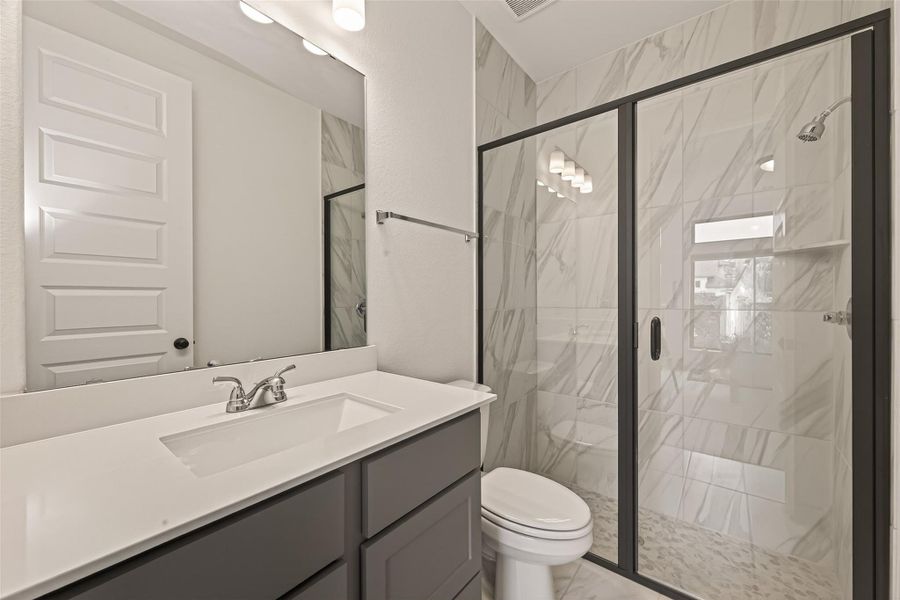 This bathroom features a modern design with a sleek white vanity, spacious mirror, and stylish gray cabinetry. It includes a glass-enclosed shower with elegant marble-patterned tiles, creating a clean and sophisticated look.