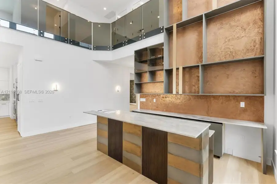 Double-height living area featuring a custom floor-to-ceiling built-in shelving unit with metallic finish backdrops, a stone-topped bar with wood and metal paneled cabinetry, light-toned wood-finish flooring, and a glass-paneled metal railing