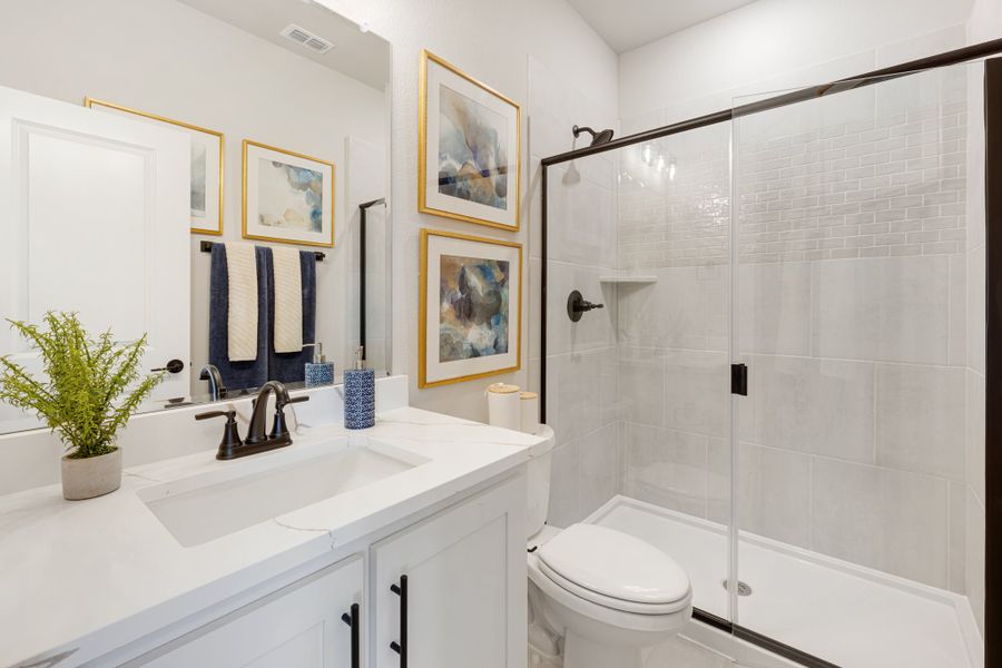 Bathroom with white vanity, glass-enclosed tile shower, toilet, and framed wall art with dark hardware accents.