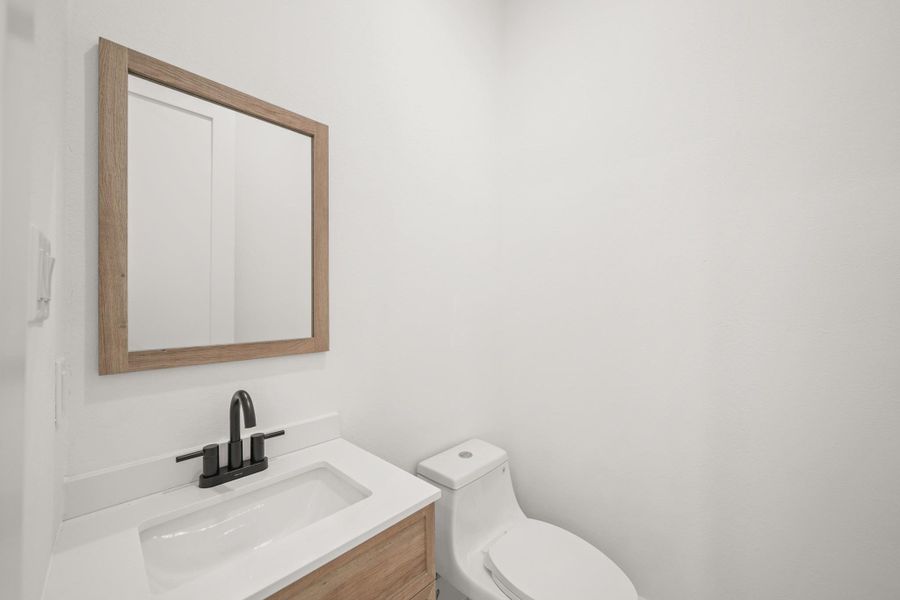 Perfect for guests, the half bath offers a sleek quartz counter, wood framed mirror and cabinetry below, a recessed light, tile floors and elegant black fixtures.