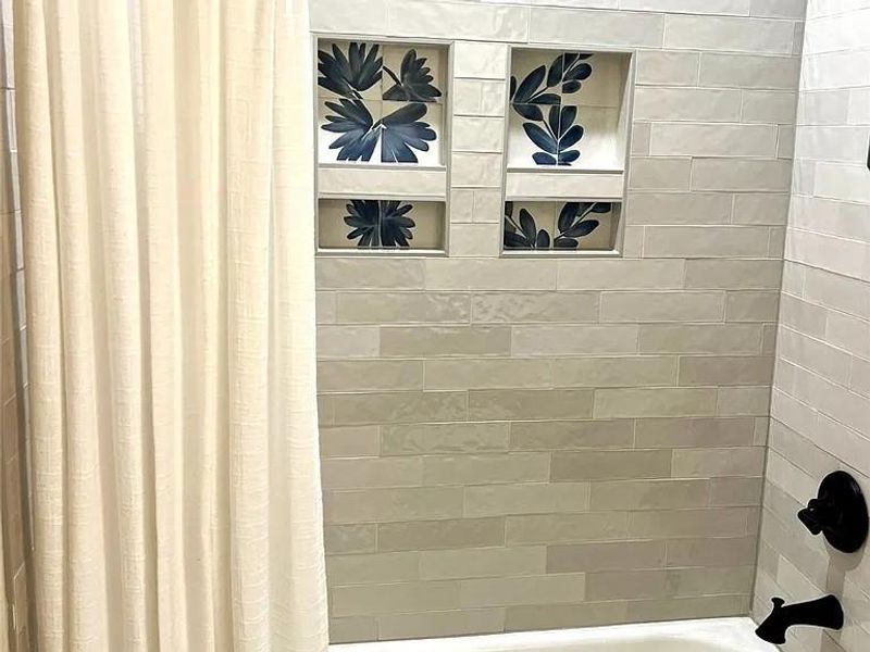 Tiled shower surround featuring integrated decorative niches with botanical accents, a built-in tub, and matte black fixtures Tiled shower surround featuring integrated decorative niches with botanical accents, a built-in tub, and matte black fixtures