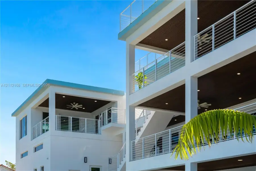 Multi-level exterior featuring stacked covered balconies, white horizontal railings, recessed lighting, and dark-toned plank ceilings Multi-level exterior featuring stacked covered balconies, white horizontal railings, recessed lighting, and dark-toned plank ceilings