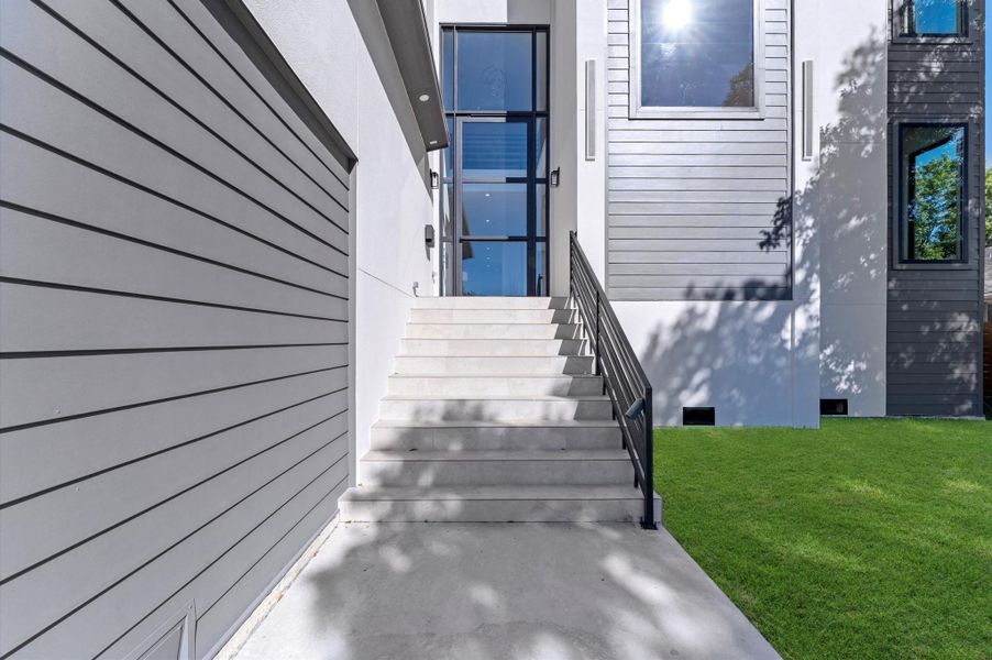 Modern home entrance with sleek lines, featuring a staircase leading to a glass front door. The exterior showcases a mix of horizontal siding and large windows, complemented by a well-maintained lawn.
