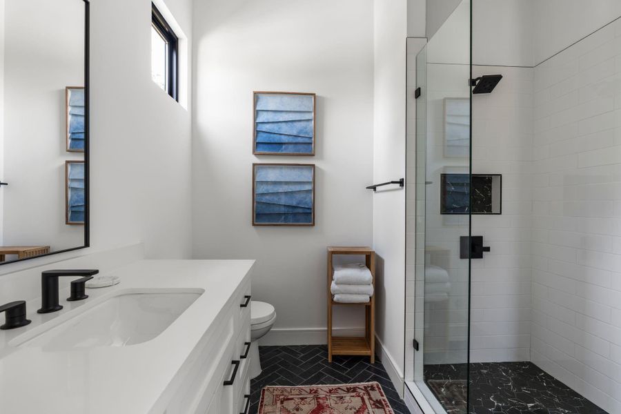 The ensuite bathroom features black herringbone tile flooring, a glass enclosed shower with white Subway tiles around, black marble shower flooring, a built-in niche, black fixtures, and a window for natural light.