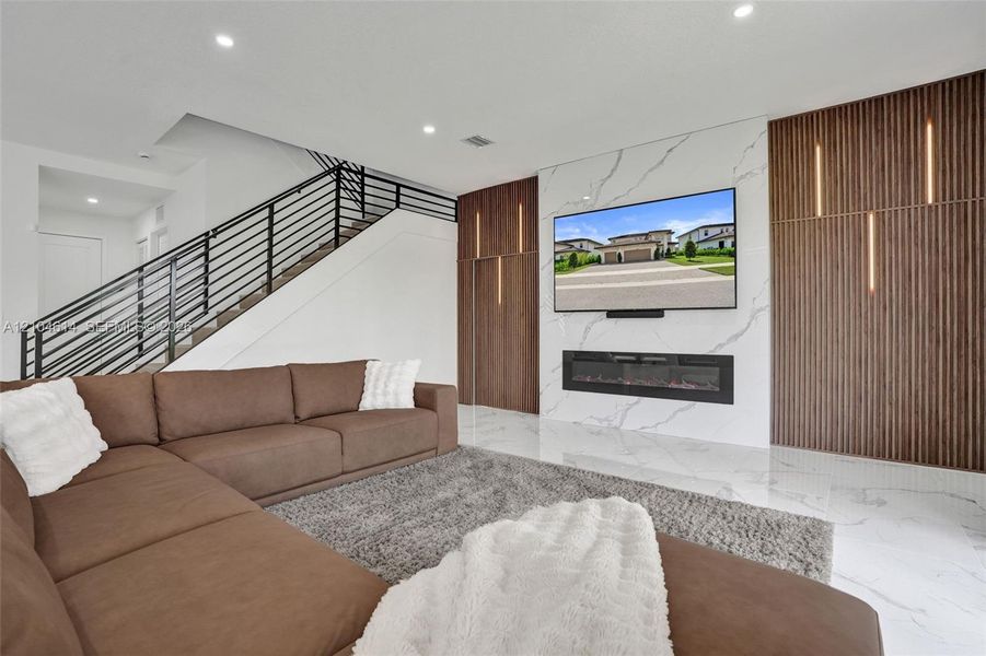 Integrated fireplace with a white marbled look accent wall, recessed lighting, wood-finish paneling with vertical lighting elements, and sleek metal stair railing