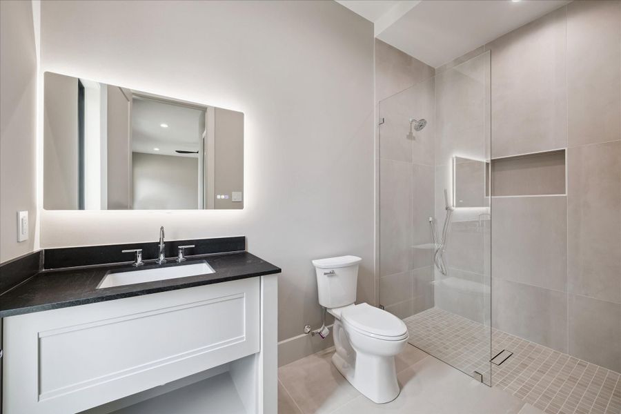 This en-suite secondary bathroom is finished with a sleek vanity topped with a quartz counter, an undermount sink, and a modern lighted vanity mirror. A spacious walk-in shower with a built-in soap inset completes the design.
