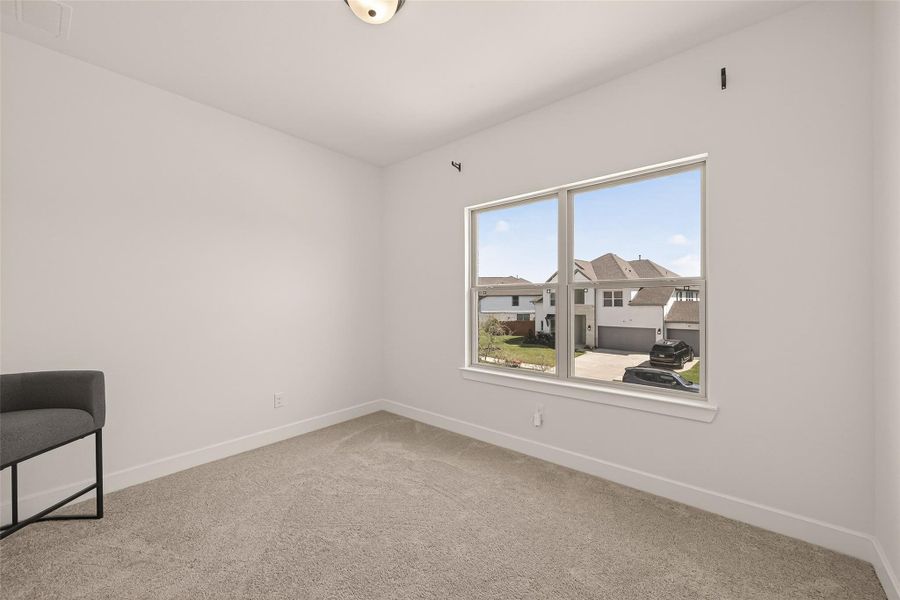 This bright bedroom features neutral carpeting and a large window with a view of a suburban street. Ideal for a home office or bedroom.