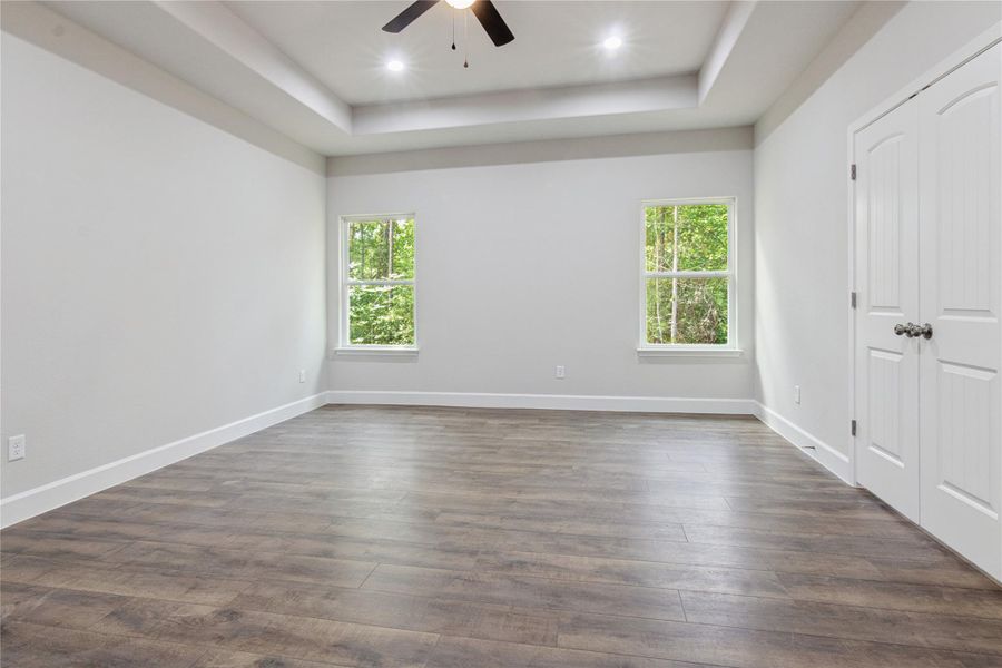 This spacious primary bedroom features elegant flooring, neutral walls, and a tray ceiling with recessed lighting and a ceiling fan. Two large windows provide natural light.