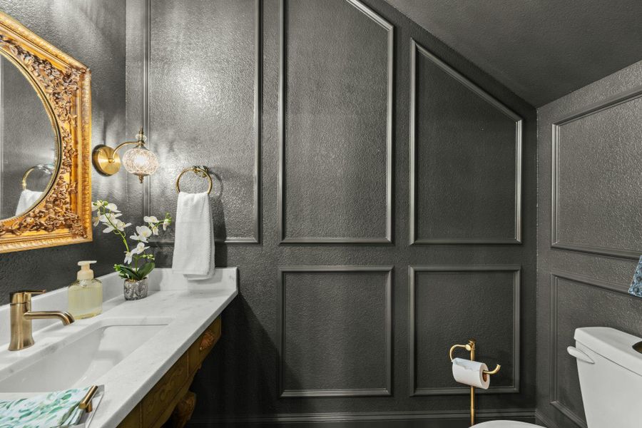 Powder room featuring detailed paneled walls, a single-sink vanity with a white countertop, and a decorative gold-framed mirror