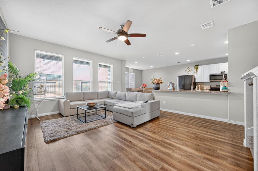 This modern living room features an open layout with sleek wood flooring and abundant natural light from large windows. It includes a comfortable sectional sofa, a ceiling fan, and a kitchen with a granite countertop, perfect for entertaining.