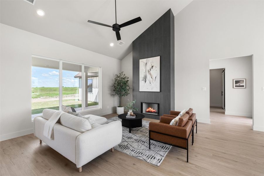 Living area with high vaulted ceiling, ceiling fan, baseboards, a large fireplace, and light wood-type flooring Living area with high vaulted ceiling, ceiling fan, baseboards, a large fireplace, and light wood-type flooring