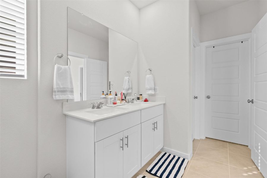 This bathroom features a modern double-sink vanity with ample storage, a large mirror, and neutral tones. It has a clean, bright aesthetic with tiled flooring and white cabinetry.