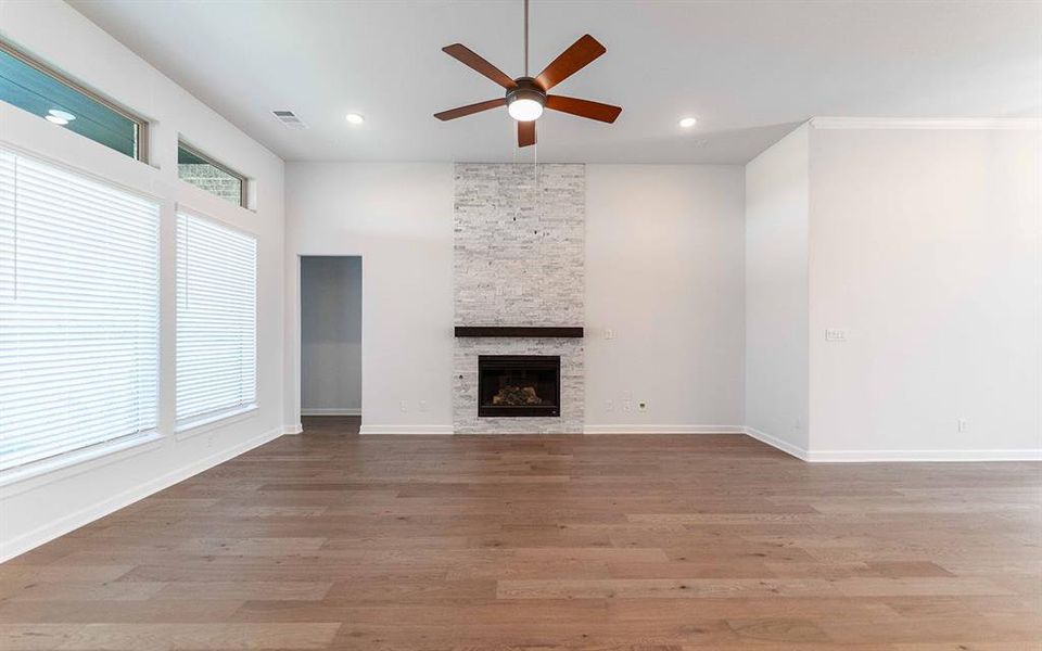 Living space featuring wood-finish flooring, a stacked stone fireplace with a dark wood mantel, and multiple windows with blinds Living space featuring wood-finish flooring, a stacked stone fireplace with a dark wood mantel, and multiple windows with blinds